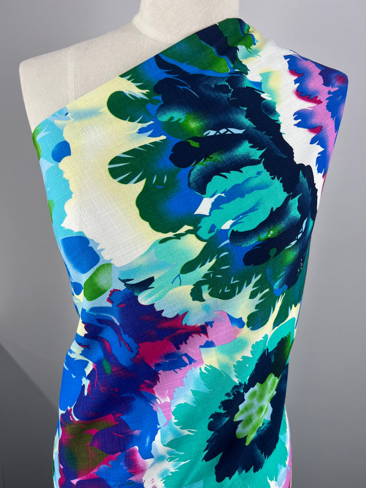 A mannequin is draped with a single shoulder fabric featuring vibrant prints in large floral patterns. The Designer Bamboo Rayon - Lantana - 147cm by Super Cheap Fabrics displays a range of colors including shades of blue, green, purple, pink, and white, forming abstract flower designs perfect for versatile clothing.