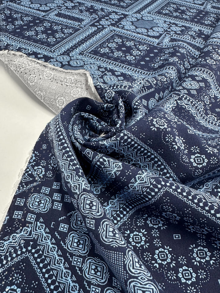 A close-up of Super Cheap Fabrics' Designer Rayon - Navy Nomad (145cm), showcasing intricate light blue geometric and floral patterns on dark blue. The versatile fabric is partially rolled to reveal its textured edge on a white background.