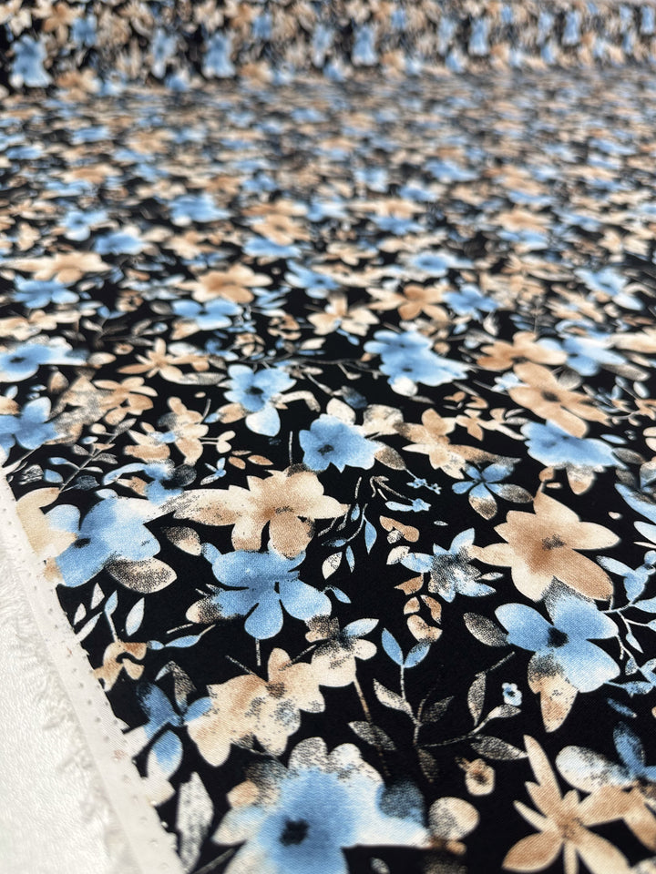 Close-up of Super Cheap Fabrics’ Designer Rayon - Dusty Petals (145cm), showcasing its lightweight rayon with a blue and beige floral pattern and green leaves on a black background. Fabric extends into the distance.