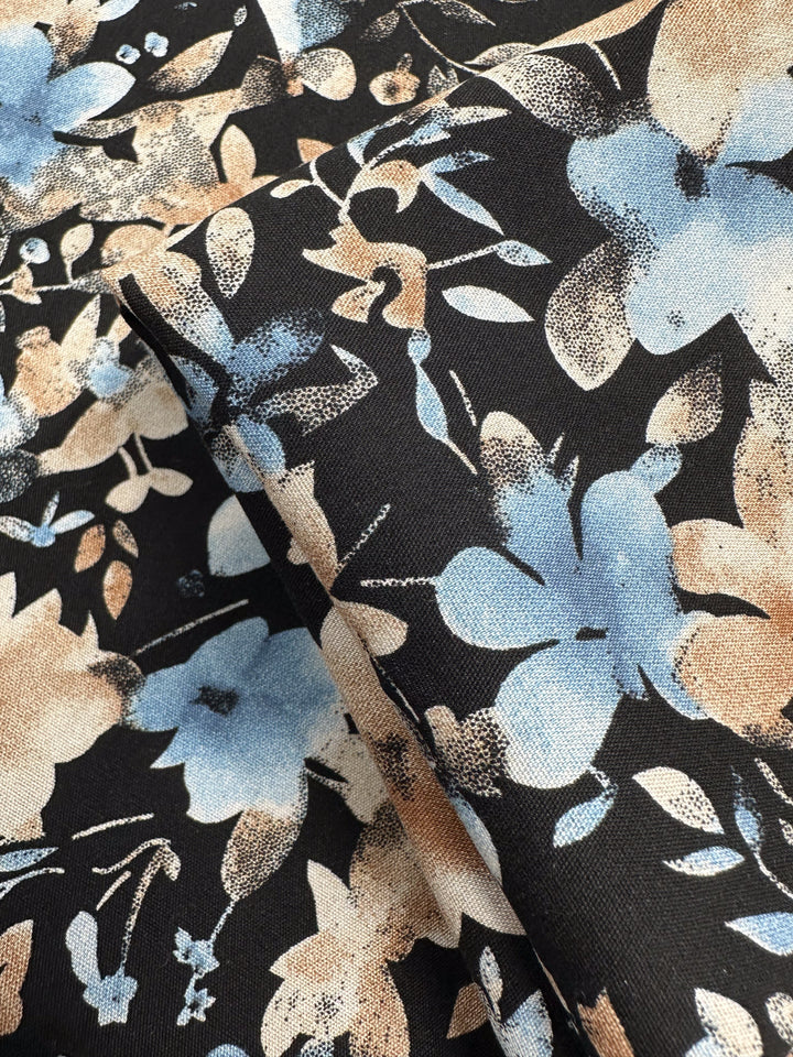 Close-up of Designer Rayon - Dusty Petals (145cm) by Super Cheap Fabrics, featuring lightweight rayon with light blue and beige floral patterns on a black background. Fabric is gently folded to display the design from various angles.