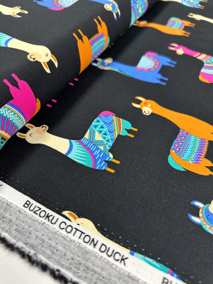 Introducing the "Cotton Duck - Lama Lama - 120cm" by Super Cheap Fabrics: a fashion-forward material showcasing colorful llamas in various poses against vibrant patterns on a black background. Ideal for home décor projects, this fabric is both stylish and versatile, with its brand label elegantly displayed along the edge.