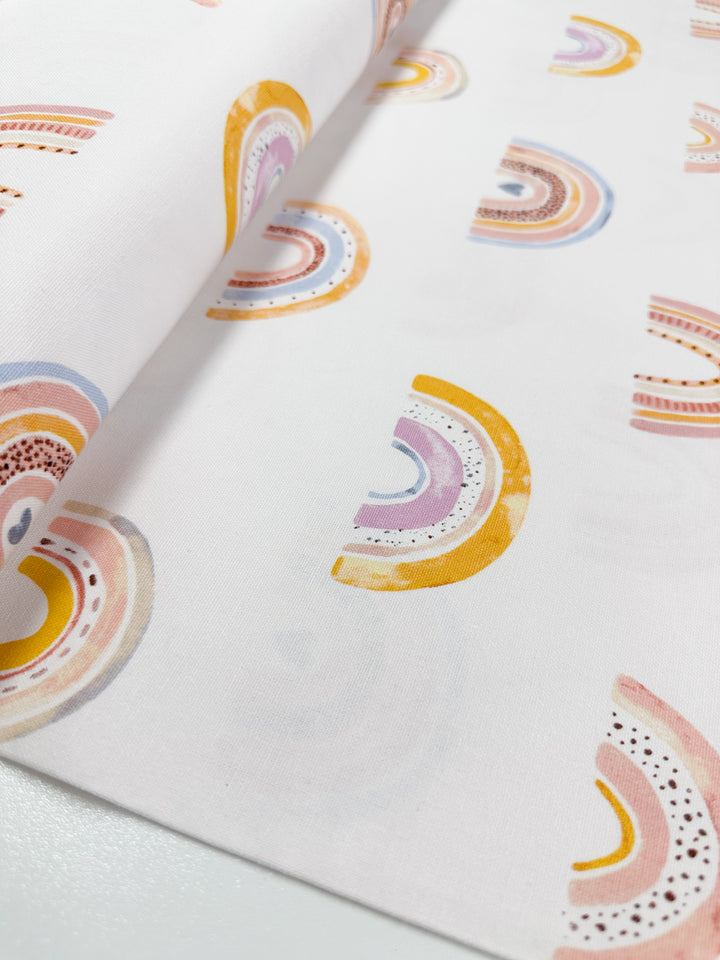 The Cotton Duck - Rainbow - 112cm from Super Cheap Fabrics showcases a pattern of vibrant, semi-circular designs in pastel shades of pink, orange, purple, and blue. Ideal for tote bags or home décor projects, the fabric is elegantly spread out on a smooth white surface.