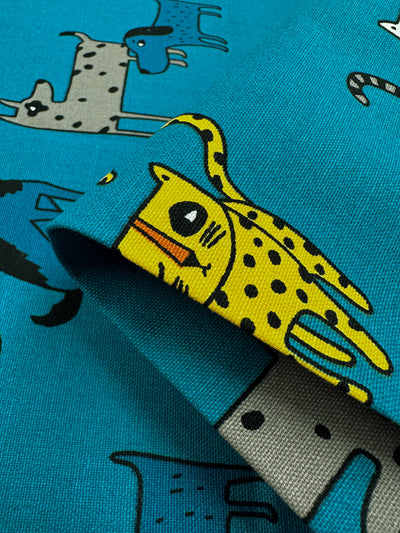 A detailed view of the Cotton Duck - Cat Dog fabric from Super Cheap Fabrics showcases its blue cotton material adorned with cheerful, cartoon animal prints in shades of yellow, gray, and blue. Ideal for home décor projects, this medium-weight fabric is folded to accentuate the whimsical yellow creature featuring spots and a distinctive eye.