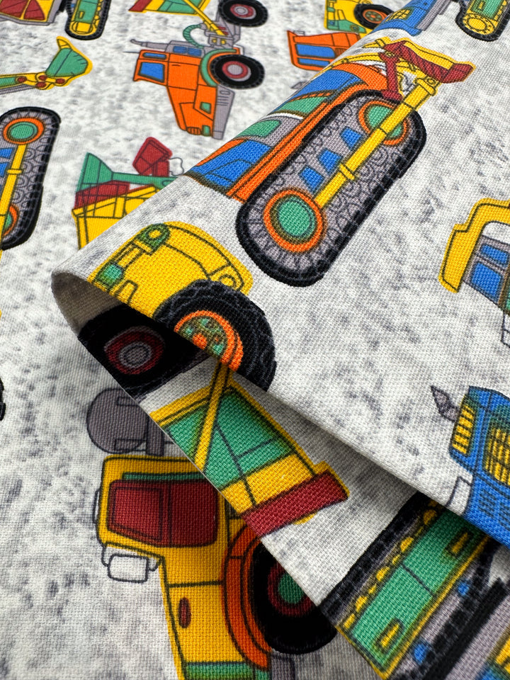 A close-up of the Super Cheap Fabrics' Cotton Duck - Construction fabric displays medium weight cotton adorned with colorful cartoon construction vehicles such as bulldozers, cranes, and trucks, set against a light gray backdrop. The fabric is folded to highlight the vibrant patterns, making it perfect for home décor projects.