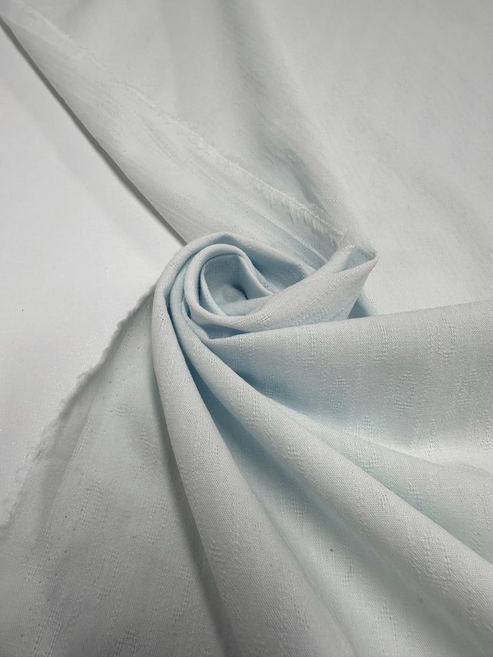 A close-up of Super Cheap Fabrics’ Textured Cotton - Ice Melt (145cm), showcasing its light blue color, subtle pattern, soft folds, gentle shadows, and lightweight texture arranged in a central swirl.
