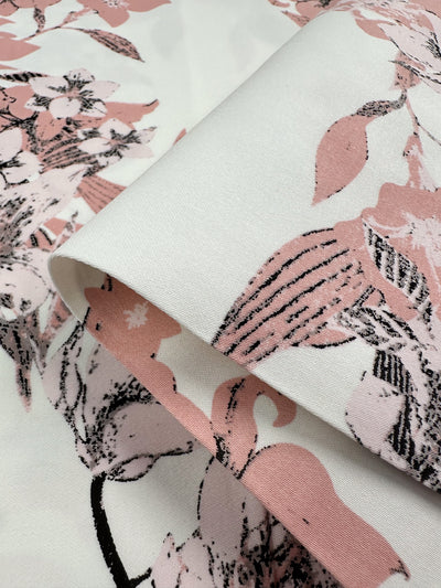 A close-up of the Cotton Sateen - Lizzy's Garden from Super Cheap Fabrics showcases a lightweight fabric with a floral pattern. The design highlights pink and black flowers and leaves on a white background, offering an elegant and soft appearance. Ideal for household décor items, the fabric is partially folded, showcasing its texture across its 128cm width.