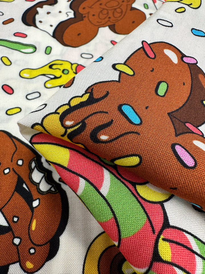 Close-up of Super Cheap Fabrics' Printed Rayon - Treat - 140cm, featuring colorful cartoon gingerbread characters and candy sprinkles. The fabric showcases layered designs in brown, yellow, green, and pink hues for a playful, festive pattern with vibrant prints.