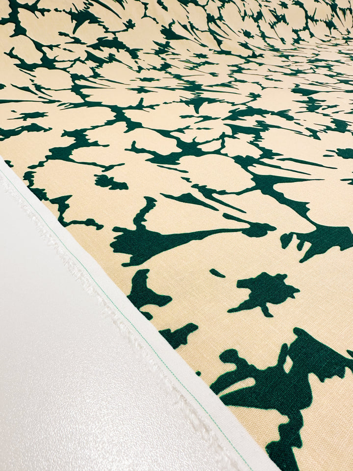 The *Printed Linen - Illusion - 135cm* by Super Cheap Fabrics showcases a beige backdrop enhanced with dark green abstract designs. The fabric's white border accentuates its versatility and intricate design allure.