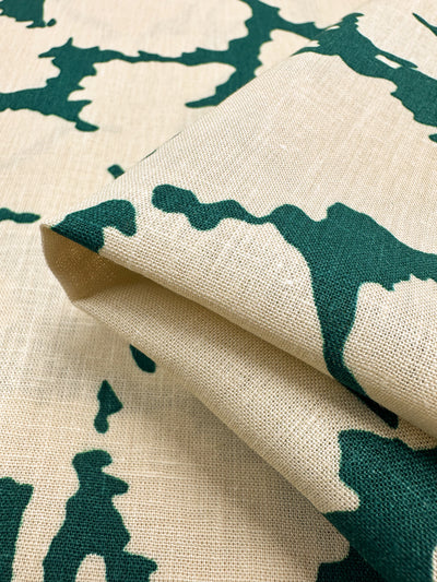 A close-up of Super Cheap Fabrics' Printed Linen - Illusion, featuring a beige background adorned with dark green abstract patterns. The 135cm versatile fabric is folded to highlight its texture and intricate designs clearly.