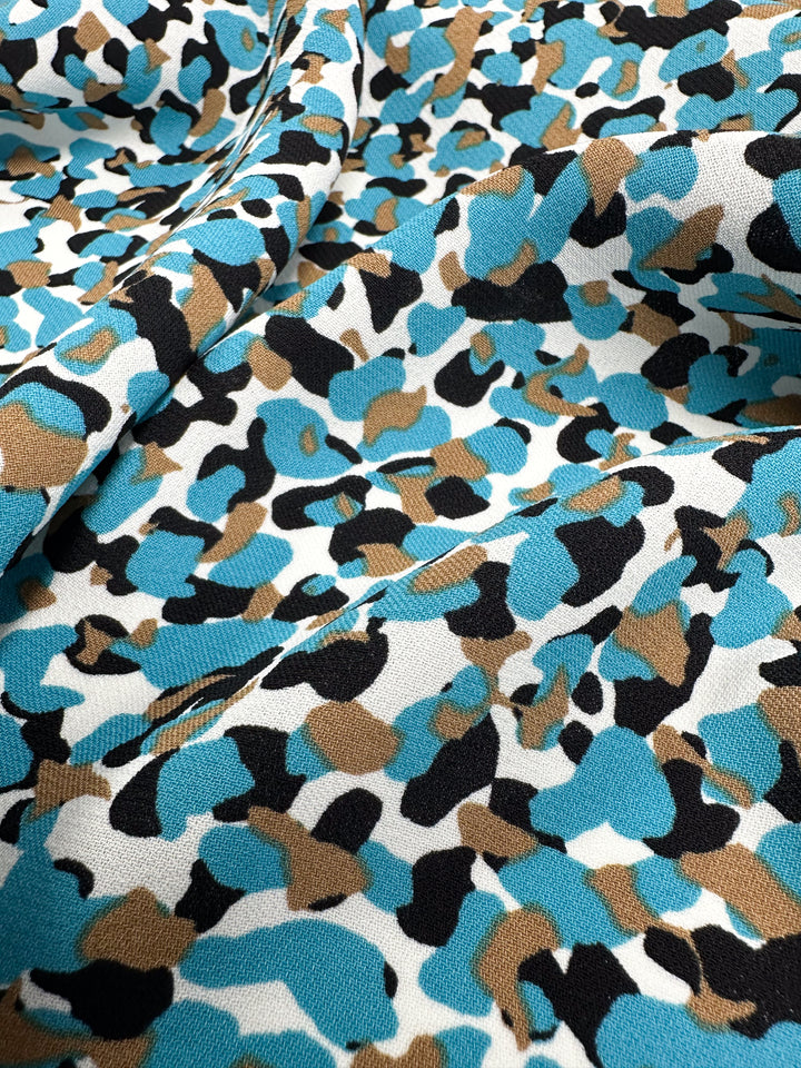 A close-up of the Viscose Crepe - Stoney fabric by Super Cheap Fabrics features a camouflage pattern blending blue, brown, black, and white hues. This luxurious material seems to be folded delicately with a textured surface, ideal for crafting versatile clothing pieces.