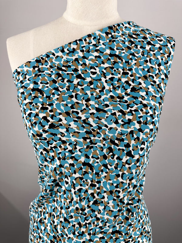 A mannequin displays a one-shoulder garment made from the Viscose Crepe - Stoney by Super Cheap Fabrics. This luxurious fabric is embellished with a colorful abstract pattern, featuring vibrant shades of blue, brown, black, and white that create a lively and dynamic print against a plain, neutral background.