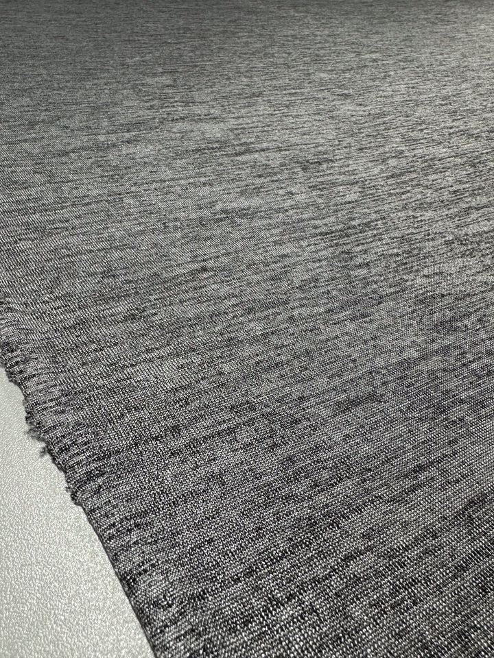 A close-up of Super Cheap Fabrics’ Textured Knit - Greyline Lamé (145cm) in dark grey, showing its textured finish and rough edge, displayed flat on a light-colored surface.