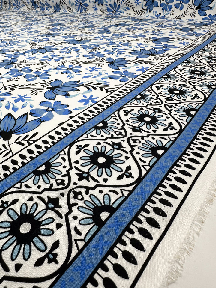 A close-up of Designer Rayon - Mosaic Valley by Super Cheap Fabrics shows blue, black, and white floral and geometric patterns with intricate borders and a slightly frayed edge, highlighting vibrant Mediterranean-inspired prints.