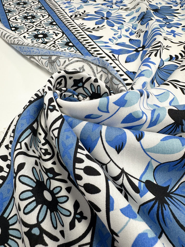 A close-up of Designer Rayon - Mosaic Valley (145cm) by Super Cheap Fabrics, showing soft rayon fabric with a decorative blue, black, and white floral pattern featuring intricate edges and bold Mediterranean-inspired blue flowers.