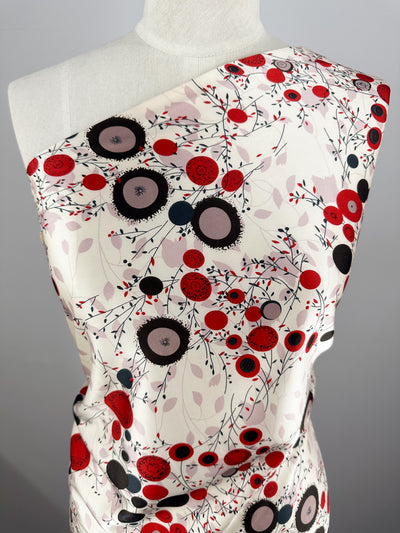 A mannequin displays the Silky Satin - Dried Flower by Super Cheap Fabrics, featuring a one-shoulder design with red, black, and pink abstract flowers and leaves on a crisp white background, creating an elegant visual on lightweight satin.