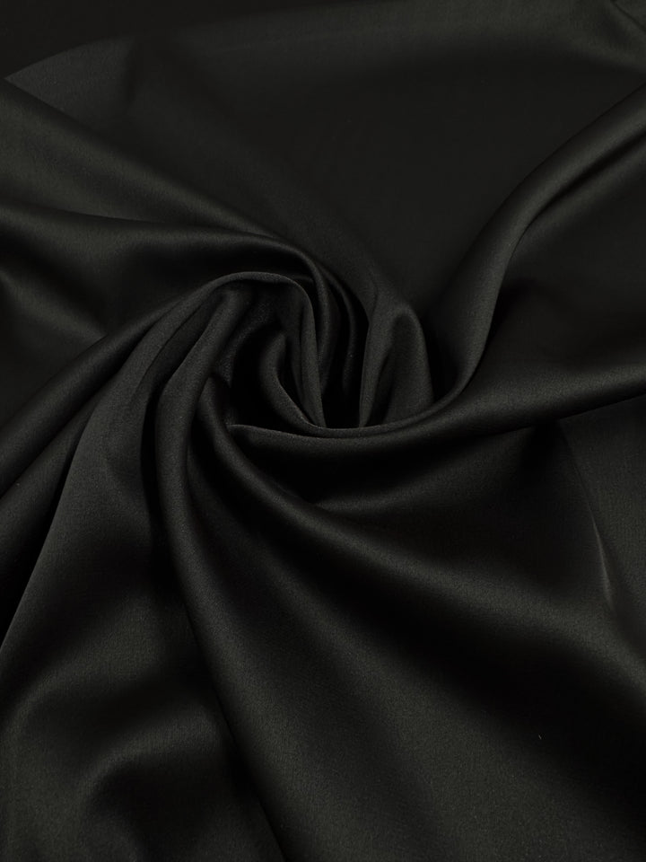 Silky Satin - Black - 150cm by Super Cheap Fabrics features a smooth, matte finish with gentle folds that create a soft, flowing pattern and subtle highlights on the fabric’s surface.