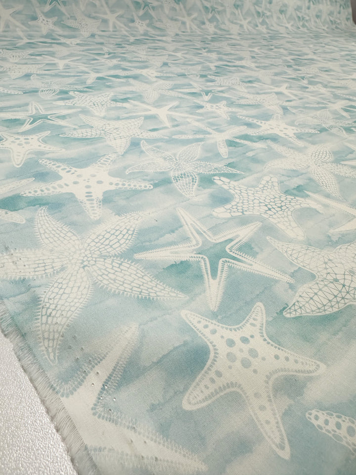 The "Under The Sea" printed cotton fabric from Super Cheap Fabrics, measuring 150cm wide, showcases a delightful design of white starfish on a light blue backdrop. Made from 100% natural cotton fibers, it captures an oceanic theme with gentle texture variations extending across the fabric.