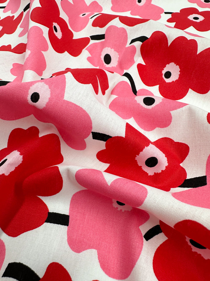 A close-up view of the "Printed Cotton - Little Miss Sunshine - 150cm" by Super Cheap Fabrics showcases its stylish design with red and pink flowers featuring black centers and white accents on a white background. Crafted from natural cotton fibers, the fabric's slight crumpling creates gentle folds and shadows, emphasizing its versatility.