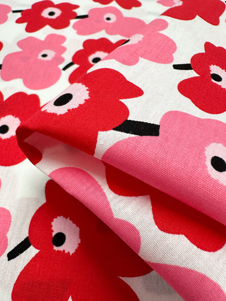 Close-up of the Printed Cotton - Little Miss Sunshine fabric by Super Cheap Fabrics, featuring a vibrant floral pattern with red and pink flowers and black stems on a white background. Crafted from natural cotton fibers, this 150cm wide fabric is folded to showcase its lively design, perfect for versatile applications.