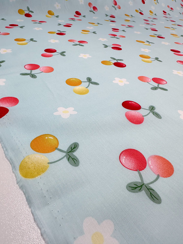 This Printed Cotton - Ripe fabric from Super Cheap Fabrics features a light blue base with a charming design of red and yellow cherries, lush green leaves, and soft white flowers. Made entirely from natural cotton fibers, it offers both aesthetic appeal and excellent breathability.