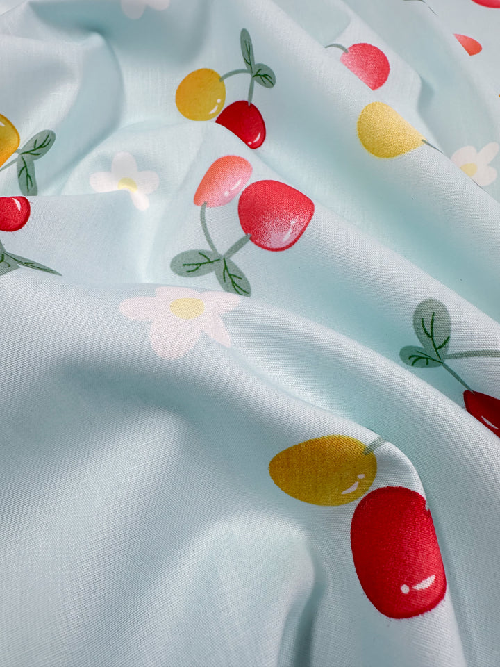 A close-up of "Printed Cotton - Ripe - 150cm" by Super Cheap Fabrics reveals a charming pattern of red and yellow cherries with green stems and white flowers. Captured in soft lighting with gentle folds, this 100% natural cotton fabric adds a delightful touch to any project.