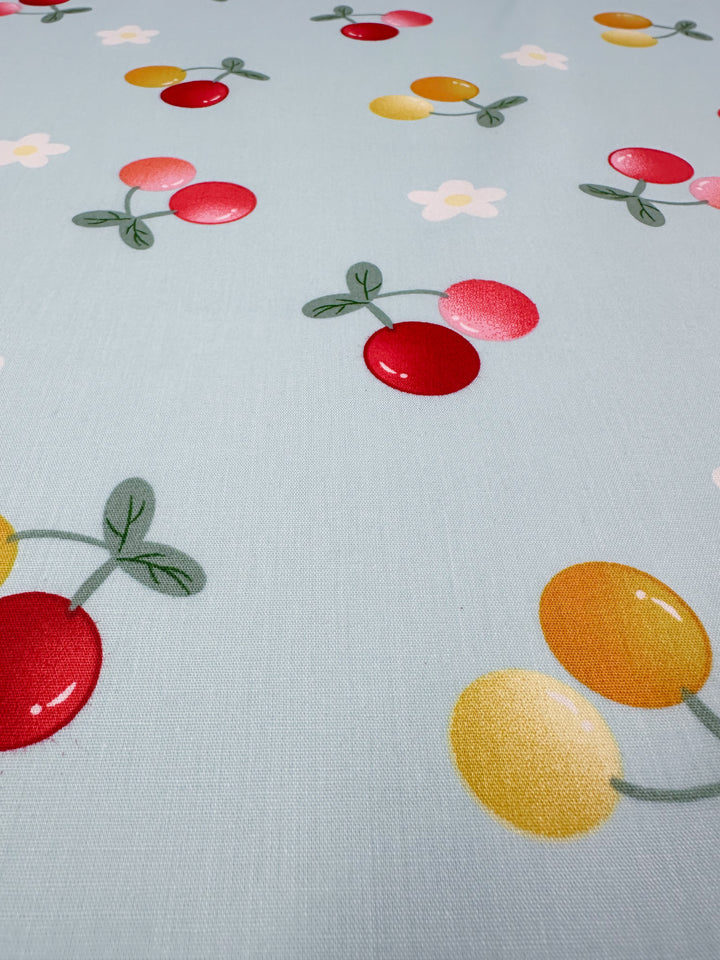 Introducing the "Printed Cotton - Ripe - 150cm" by Super Cheap Fabrics, a delightful fabric featuring a light blue background adorned with bright red and yellow cherries, some accompanied by green leaves, and accented with small white flowers. Made from 100% natural cotton fibers, this fabric is the perfect choice for adding a cheerful touch to your projects.