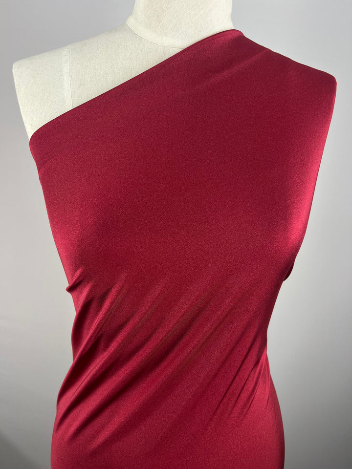 A mannequin is adorned in an elegant one-shoulder dress made of Super Cheap Fabrics' Nylon Lycra in the rich, deep hue of Rumba Red. The smooth fabric, measuring 160cm wide, is slightly gathered to create subtle wrinkles that enhance the dress's texture and sophistication. The background is a plain, neutral color.