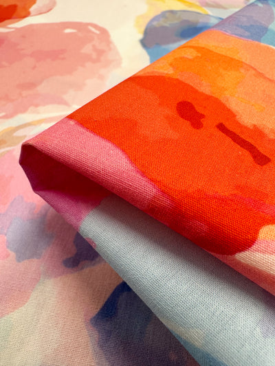 A close-up of folded Printed Cotton - Bubble - 150cm from Super Cheap Fabrics showcases a vibrant abstract pattern in shades of red, orange, pink, and blue. The natural cotton fibers provide a colorful and artistic display against a similarly patterned background, making it ideal for clothing and household items.