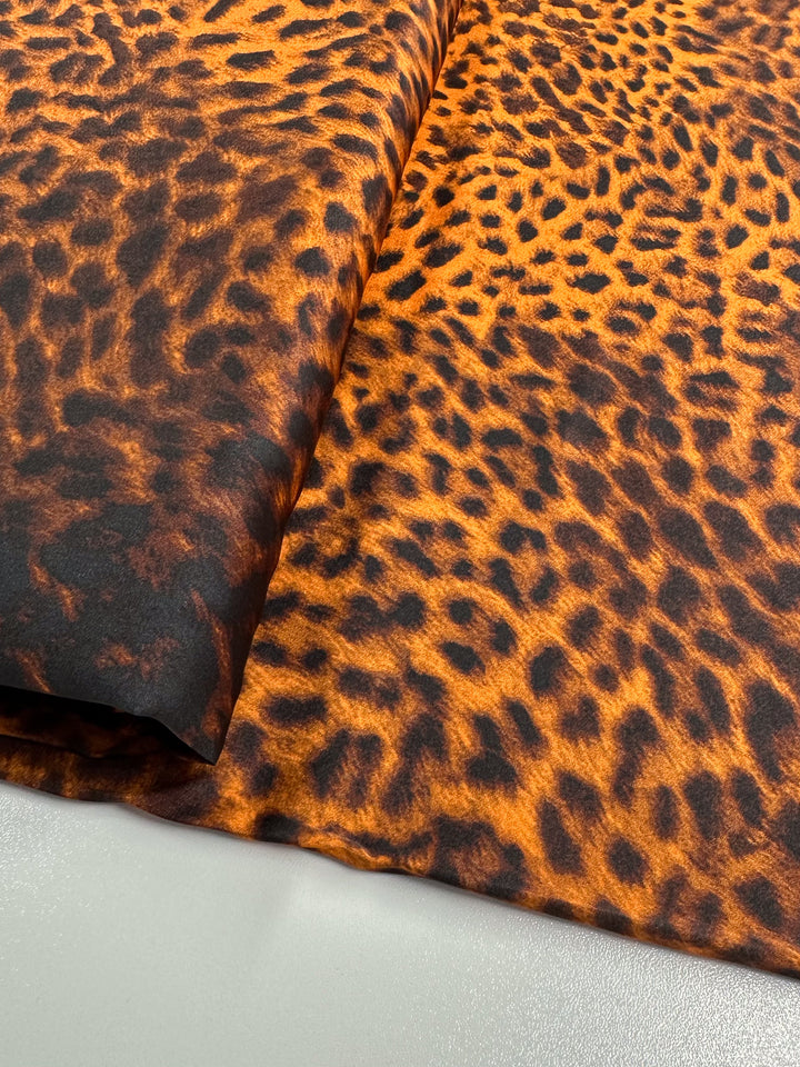 Close-up of folded Silky Satin - Namer - 145cm from Super Cheap Fabrics, showcasing a vibrant leopard print pattern in shades of orange and black. This lightweight satin, with a slight sheen, is elegantly displayed on a light surface.