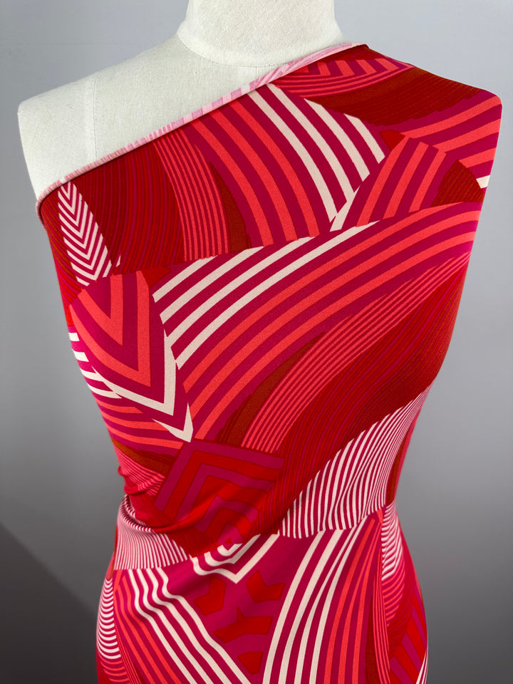 A close-up of a mannequin adorned with a vibrant, one-shoulder garment made from medium-weight Printed Lycra - Wonka - 150cm fabric by Super Cheap Fabrics and featuring a geometric pattern. The fabric showcases bold, wavy stripes in shades of red, pink, and white, creating a dynamic and eye-catching design.