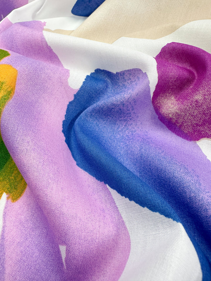 Close-up of Super Cheap Fabrics' Printed Cotton - Petals, showcasing a vibrant design with abstract, blended patches of purple, blue, pink, and green. The smooth yet slightly wavy texture crafted from natural cotton fibers adds a dynamic visual effect.