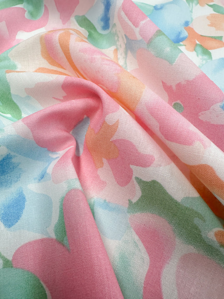 A close-up view of the "Printed Cotton - Blossoms - 150cm" by Super Cheap Fabrics reveals a colorful floral pattern. The design prominently features pink flowers accented with touches of blue, green, and orange for a soft yet vibrant appearance. Crafted from natural cotton fibers, this slightly gathered fabric boasts a textured finish ideal for various applications.