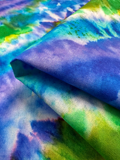 A close-up of the Printed Cotton - Lizard fabric from Super Cheap Fabrics reveals its vibrant tie-dye design, crafted from natural cotton fibers. This material showcases swirling patterns in blue, green, yellow, and purple hues. The fabric is folded to highlight its colorful design and soft texture.