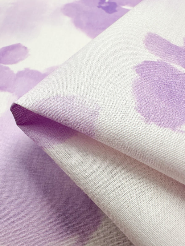 Close-up of a folded Super Cheap Fabrics' Printed Cotton - Bellflower, featuring a soft, abstract watercolor pattern in light purple and white. The natural cotton fiber texture is visible, with pastel hues that offer a gentle and calming appearance ideal for home decor.