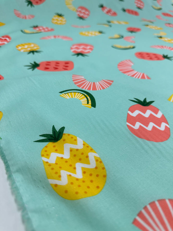 This fabric, branded as Super Cheap Fabrics' "Printed Cotton - Pina Coalda - 150cm," is crafted from natural cotton fibers. It features a pastel mint green background decorated with vibrant fruit patterns, including playful pineapples in yellow and pink, strawberries, and abstract citrus slices with zigzag and dotted designs. It's ideal for both clothing and household items.