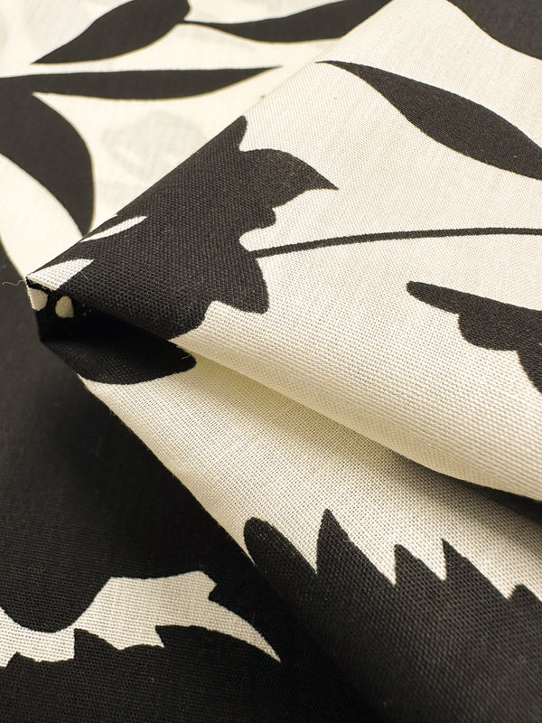 Close-up of the "Visage" printed cotton fabric, featuring a striking black-and-white leaf pattern. The natural cotton fibers from Super Cheap Fabrics appear folded, showcasing the contrast between dark and light areas while highlighting the texture and woven detail of this 150cm wide product.