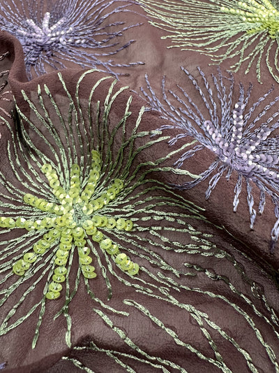 A close-up of the Pure Printed Silk - Fireworks from Super Cheap Fabrics highlights detailed embroidery, where sequins and threads create green and purple floral designs against a brown background. The embroidered flowers, with petal-like patterns that radiate outward, present a vibrant and textured look.