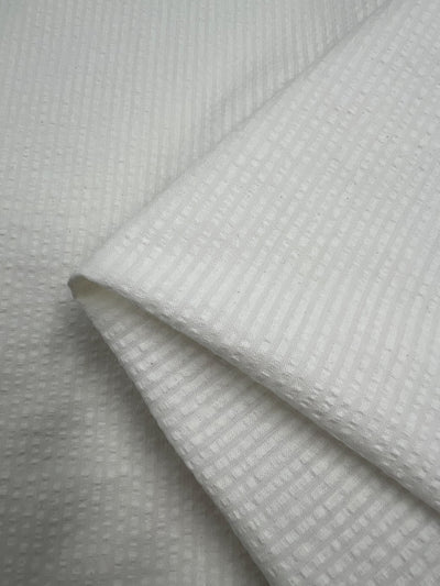 A close-up photo of neatly folded Seersucker Cotton - Off White - 155cm fabric with a subtle textured pattern of small squares and ridges. The medium weight cotton fabric from Super Cheap Fabrics appears soft and clean, with the pattern creating a visually interesting detail.