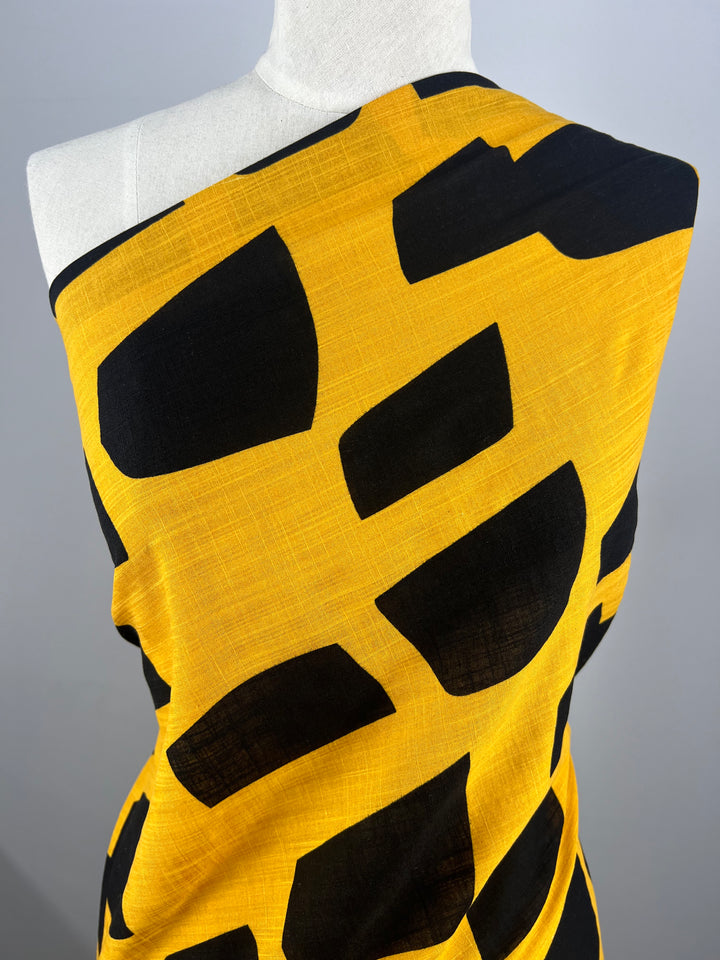 A mannequin is draped with a vivid yellow Bamboo Rayon - Bedrock fabric from Super Cheap Fabrics. This beginner-friendly, 152cm wide fabric features large, irregular black geometric shapes. The lightweight material is wrapped asymmetrically over one shoulder, highlighting its bold and striking pattern against a plain grey background.