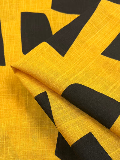 A close-up image of yellow Bamboo Rayon - Bedrock - 152cm fabric from Super Cheap Fabrics, featuring large, abstract black geometric shapes. The lightweight material is slightly folded, showcasing the texture and woven pattern of this fabric.