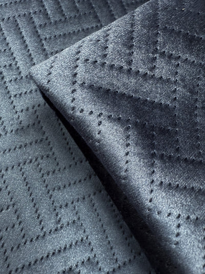 Close-up of Super Cheap Fabrics' Embossed Velvet - Oceana (150cm) in dark grey, showing a subtle zigzag and dotted pattern. The folded corner highlights this upholstery fabric's rich texture and sophisticated design.