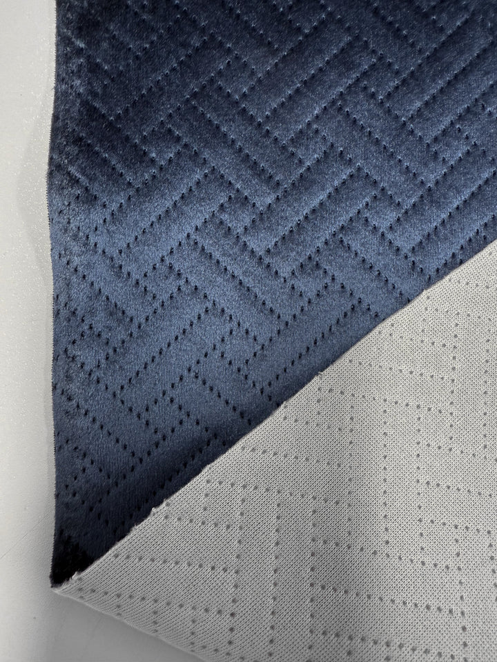 A close-up of Super Cheap Fabrics’ Embossed Velvet - Oceana (150cm), a heavy blue velvet with a geometric zigzag pattern, partially folded to show its light gray underside with a similar design.