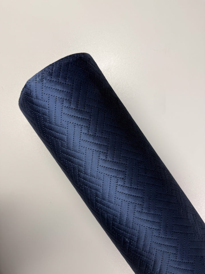 A dark blue cylinder of Embossed Velvet - Oceana (150cm) by Super Cheap Fabrics, featuring a geometric woven pattern, lies diagonally on a plain light surface.