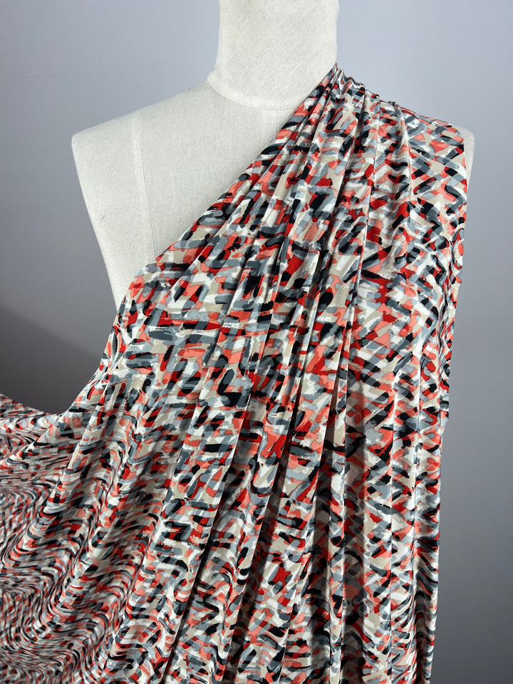 A mannequin is draped in Super Cheap Fabrics' Printed Lycra - Stroke, a medium-weight fabric measuring 150cm wide, featuring a colorful abstract pattern with shades of red, black, gray, and white. The fabric is skillfully arranged to cover one shoulder and cascades down to create gathered folds that showcase its textured design. The backdrop is plain gray.