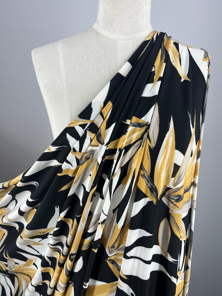 A mannequin draped in Super Cheap Fabrics' Printed Lycra - Bamboo, a medium-weight Polyester/Spandex fabric, showcases a bold botanical design featuring large leaves in shades of gold, white, and black swirls against a dark background. The material is styled asymmetrically over one shoulder.