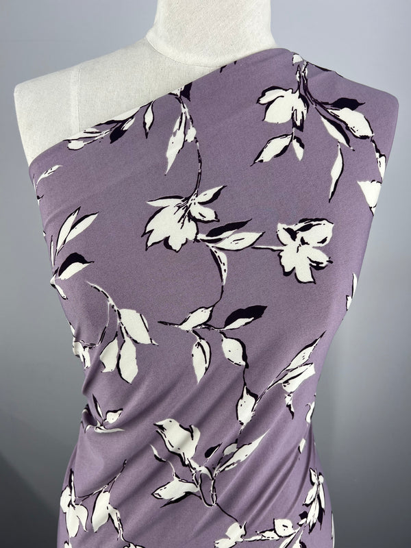 A mannequin draped in Printed Lycra - Dreamal from Super Cheap Fabrics, showcasing an intricate off-white and dark purple floral pattern. The medium-weight fabric is styled diagonally, covering one shoulder and leaving the other bare, highlighting the design and smooth texture of this 150cm material.