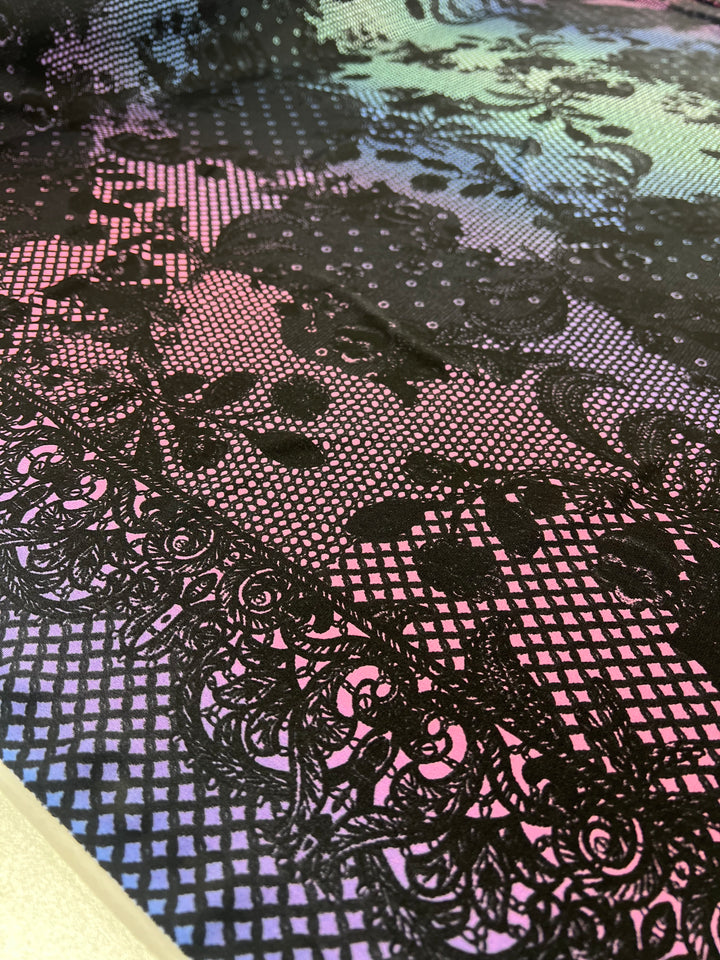 Close-up of the Super Cheap Fabrics' luxurious Printed Lycra - Dreamal - 150cm, featuring an intricate black lace pattern over a shimmering gradient background. The gradient transitions from pink to blue, accentuating the intricate floral and geometric lace designs on this medium-weight Polyester/Spandex fabric.