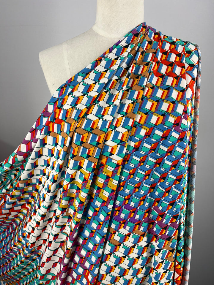 A mannequin draped in **Printed Lycra - Rubex - 150cm** by **Super Cheap Fabrics**, showcasing a vibrant, multicolored geometric pattern featuring interlocking cubes in shades of blue, orange, red, green, purple, and white. The versatile medium-weight fabric creates a visually dynamic design against a neutral gray background.