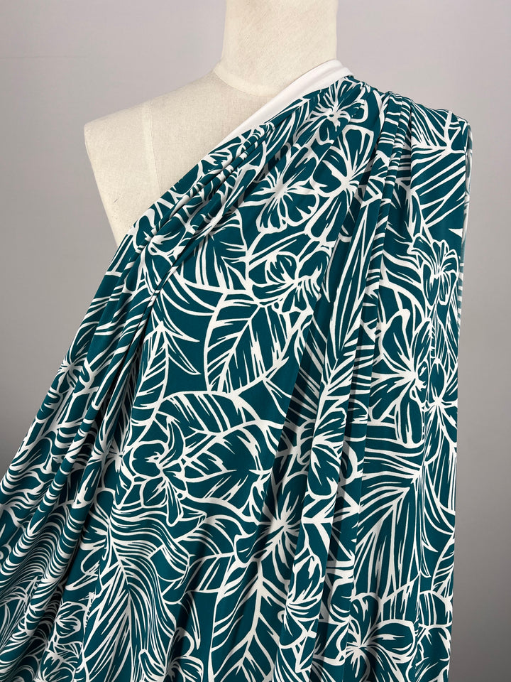 A mannequin draped in Super Cheap Fabrics' Printed Lycra - Jasmint - 150cm, featuring a bold green and white floral pattern. The fabric elegantly covers one shoulder and cascades down, showcasing the large floral and leaf designs against a plain gray background that highlights the intricate details of this two-way stretch material.