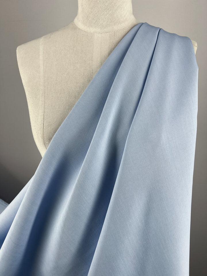 A mannequin draped with the Super Cheap Fabrics' Suiting - Clear Sky (142cm) across its chest and shoulder is set against a plain gray background. The 97% cotton material appears soft, smooth, and medium weight, beautifully showcasing its light and airy texture.